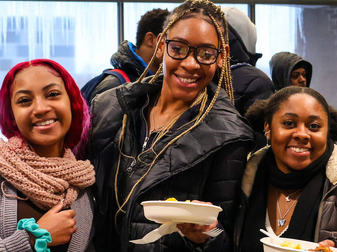 Three students smile while holding plates of food in a brightly lit indoor space.